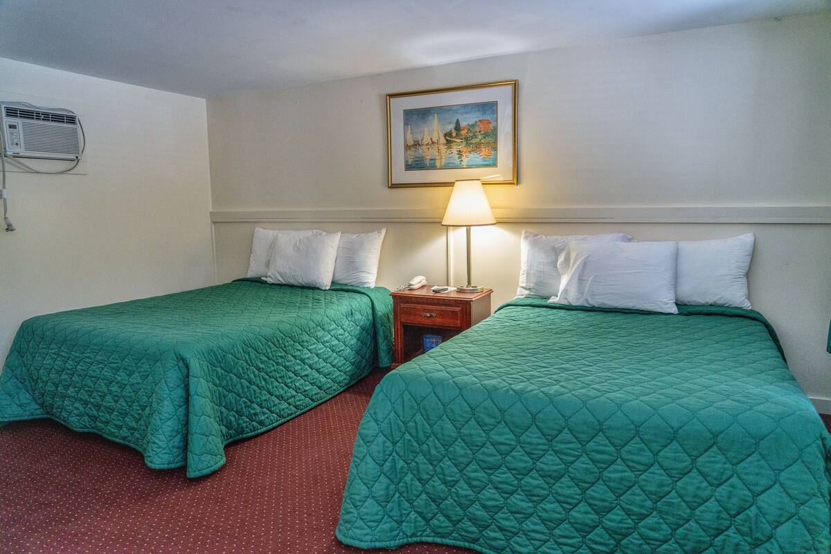 A hotel room with two beds, each with a green quilted bedspread and white pillows. There is a nightstand with a lamp between the beds. A painting hangs on the wall above the beds.