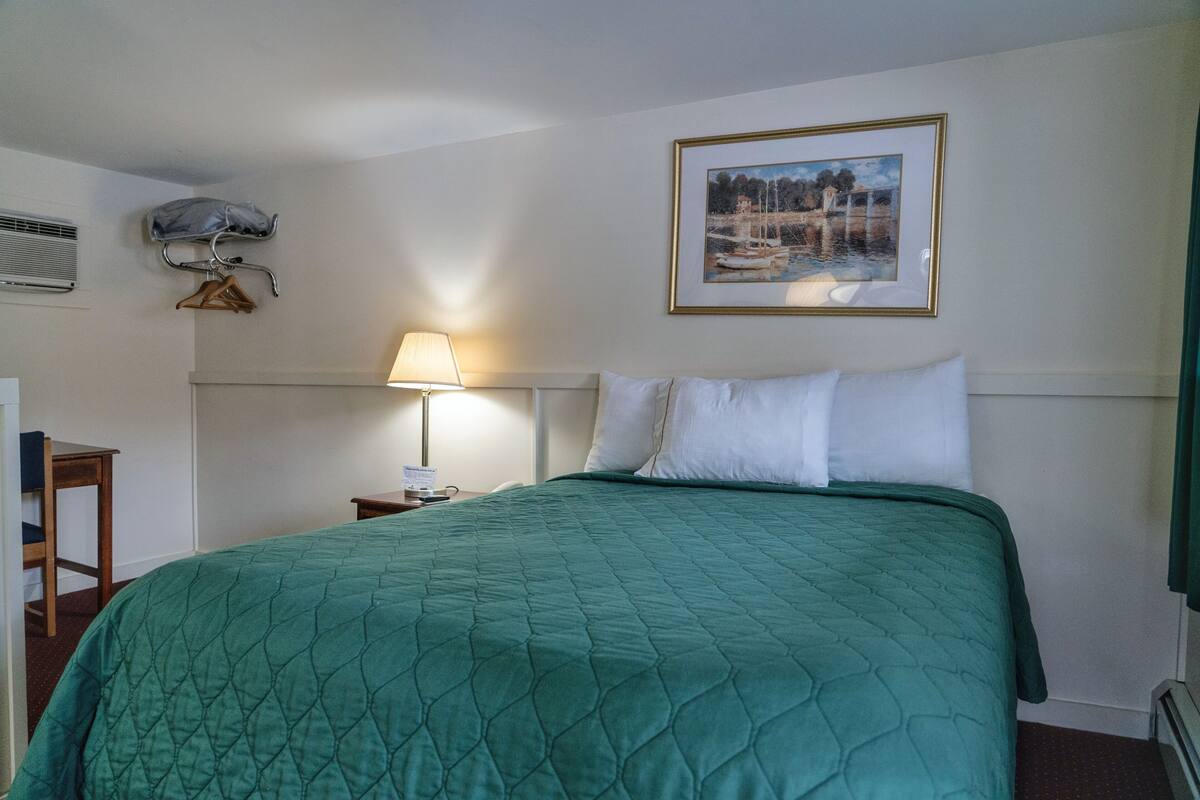 A neatly made bed with a quilted green bedspread, a bedside lamp, and a framed picture on the wall.