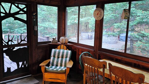 Ein gemütliches Blockhaus-Interieur mit einem Holzespeisetisch, zwei Stühlen, einem gepolsterten Sessel, einem großen Fenster mit Blick auf einen Wald und einer Wanduhr.