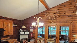 Ein rustikales Holzinterieur mit einem für sechs Personen gedeckten Esstisch, umgeben von Stühlen im Vintage-Stil. Eine Chandelier hängt von der Decke, und ein großes Fenster bietet Blick ins Freie.