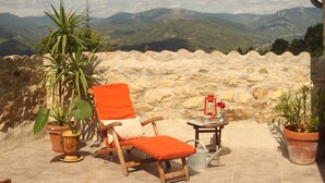Una terraza con una silla de madera y cojín naranja, plantas en macetas y un farol sobre una mesa.