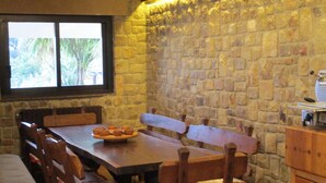 A stone-walled dining area with a long wooden table and benches, a window with a view of greenery, and a small kitchen area with a coffee machine. - Luxury Villas in Sicily with pool (Cefalù)