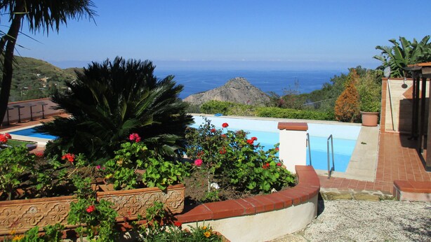 A pool area with a view of the ocean, surrounded by lush greenery and a stone wall. - Luxury Villas in Sicily with pool (Cefalù)