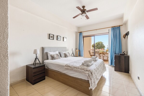 A bedroom with a bed, bedside table, lamp, ceiling fan, and a view of the outdoors.