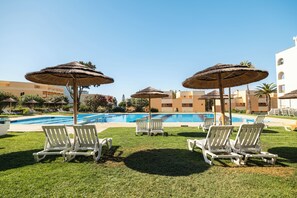 A poolside area with lounge chairs and thatched umbrellas, surrounded by buildings and greenery.
