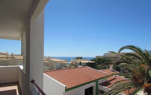 A balcony with a red railing, a white wall, and a view of the sea and other buildings.