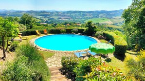 A circular swimming pool with a stone border, surrounded by lush greenery and a scenic view of rolling hills. - Stone Casale Apartment with Pool & Terrace: Sibillini to Sea View (Penna San Giovanni)