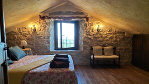 A cozy attic bedroom with a sloped ceiling, a bed with floral bedding, a chair with a cushion, and a window with a view. - Stone Casale Apartment with Pool & Terrace: Sibillini to Sea View (Penna San Giovanni)
