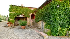 Un edificio rústico con fachada de piedra, muros cubiertos de hiedra, entrada de adoquines y un pequeño jardín con plantas en macetas.
