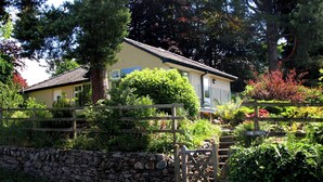 A house surrounded by a stone wall and wooden fence, with lush greenery and trees in the background. - Lovely 3 bedroom bungalow in Keswick with gardens and ample off road parking (Keswick)