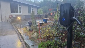 A residential area with a house, a garden with various plants, and a black electric vehicle charging station. - Lovely 3 bedroom bungalow in Keswick with gardens and ample off road parking (Keswick)