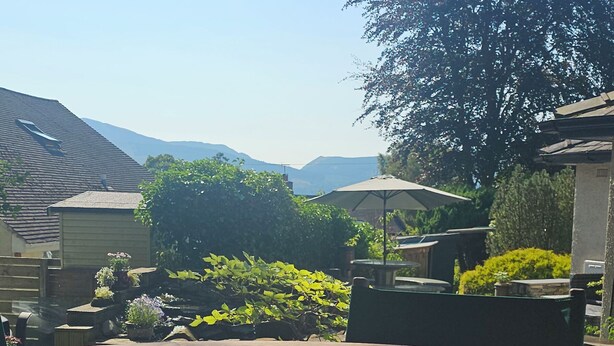 A wooden deck with a circular table, surrounded by potted plants and a large tree. - Lovely 3 bedroom bungalow in Keswick with gardens and ample off road parking (Keswick)