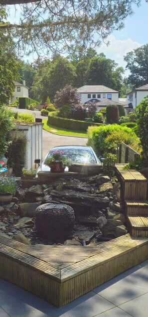 Un quartier résidentiel avec une voiture garée dans une allée, entouré de jardins bien entretenus et d’un petit étang.