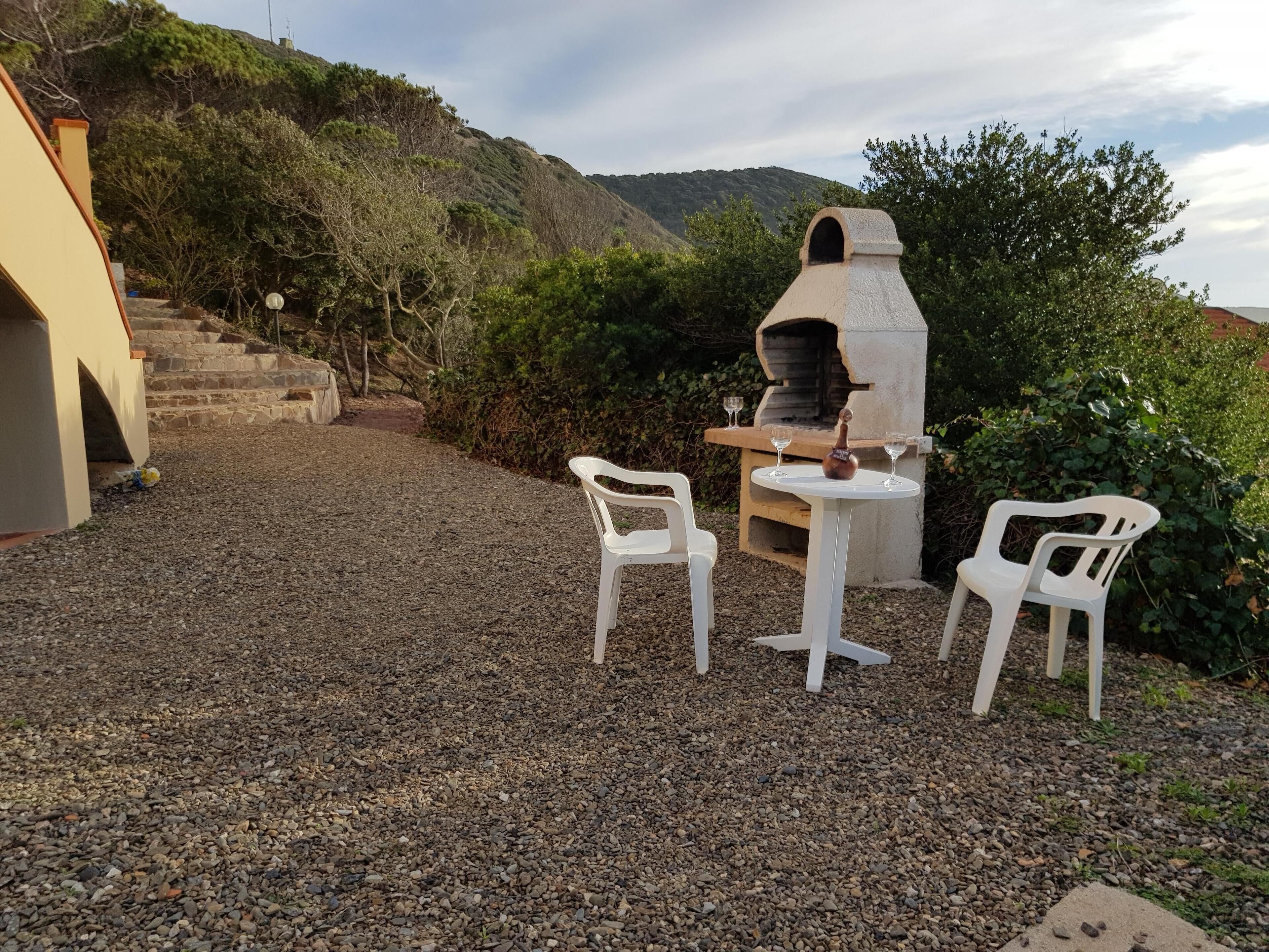 An outdoor patio with a stone table and two white plastic chairs, a stone oven, and a stone staircase leading up a hill.