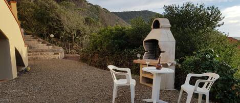 An outdoor patio with a stone table and two white plastic chairs, a stone oven, and a stone staircase leading up a hill.