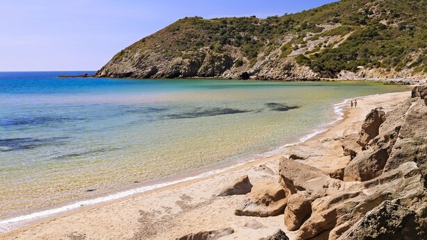 A beach with clear water, rocky terrain, and a hilly backdrop.