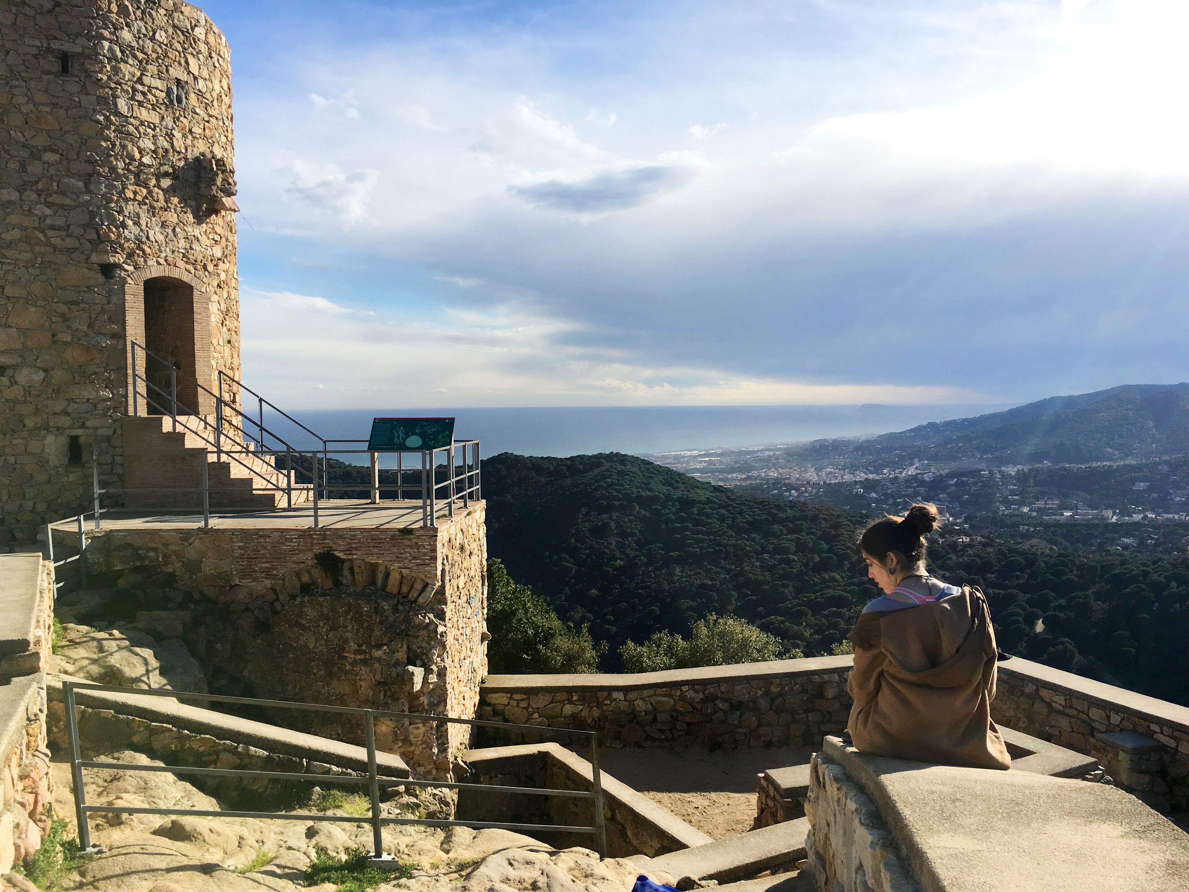 A stone tower with a viewing platform and a sign, overlooking a valley and distant city.