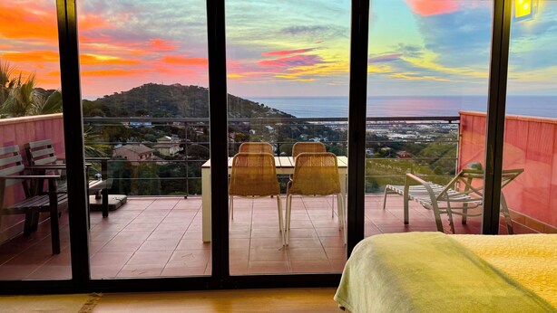 A balcony with a view of a sunset over a coastal landscape, furnished with wicker chairs and a table. - In Cabrils (Barcelona at 20 km). Apartment with sea view, pool and very quiet (Cabrils)