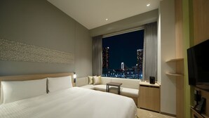 Habitación de hotel con una cama grande, un sofá, una mesita, un televisor y vistas a la ciudad de noche.
