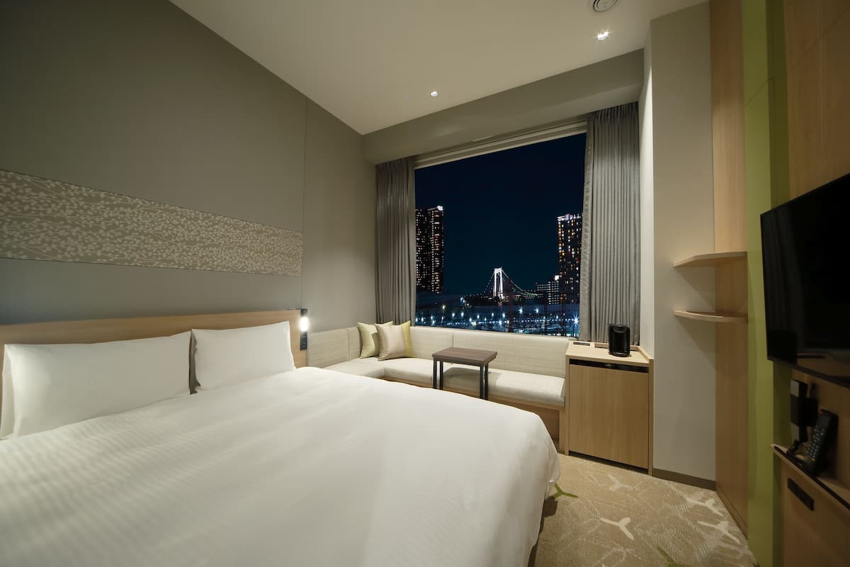 A hotel room with a large bed, a sofa, a small table, a TV, and a view of the city at night.