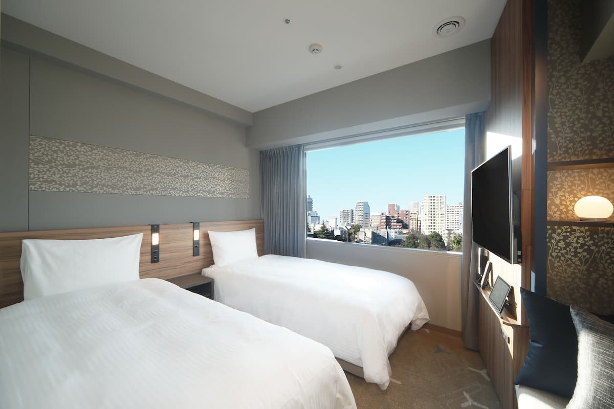 A hotel room with two beds, a TV, and a large window with a city view.