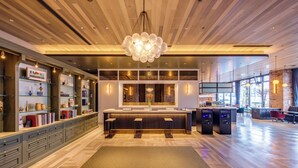 Un moderno vestíbulo de hotel con una zona de bar central, techo de madera y estanterías de vidrio con libros y objetos decorativos.