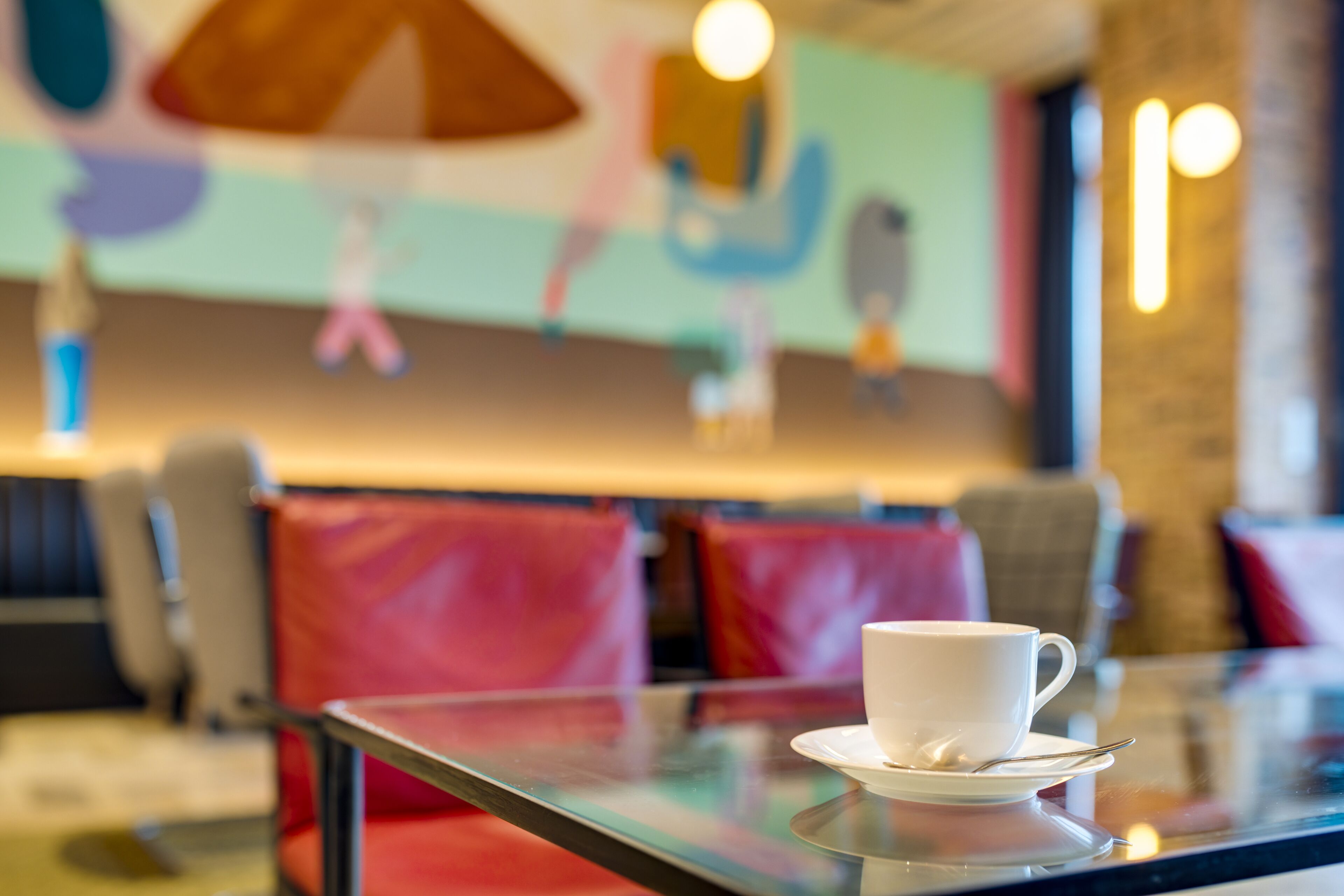 A white cup on a glass table with a saucer, set in a modern interior with red cushioned seating and a colorful mural on the wall.