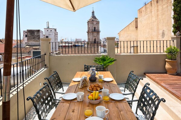 Mesa de madera preparada para el desayuno con cruasanes, fruta y tazas de café en un balcón con vistas a una ciudad que incluye una torre destacada.