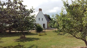 A white building with a steep roof and a chimney, surrounded by lush green trees and grass.