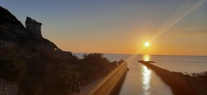 Atardecer costero con un castillo en una colina, un camino que lleva al mar y gente caminando.
