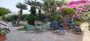 Un patio con mobiliario exterior, incluyendo una mesa y sillas, rodeado de abundante vegetación y plantas florecientes.