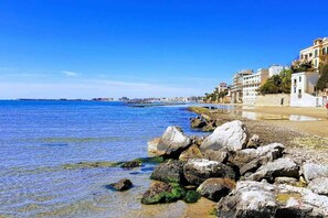 A coastal scene with clear blue water, rocky shoreline, and buildings along the beach.