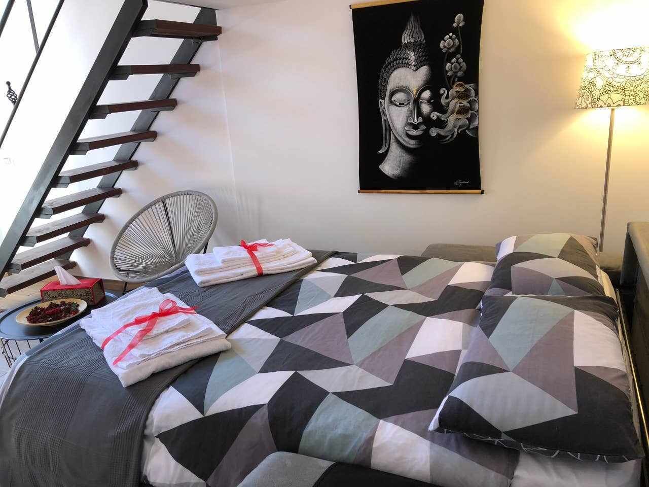 A neatly made bed with a geometric-patterned bedspread, a towel with a red ribbon, a fan, and a Buddha painting on the wall.
