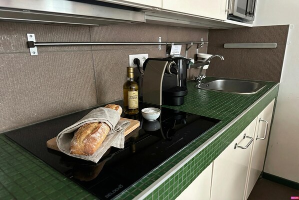 A kitchen with a green tiled countertop, a black induction cooktop, a coffee maker, a bottle of olive oil, a bowl, and a loaf of bread on a cutting board.