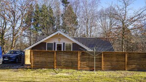 A small, single-story wooden cabin with a sloped roof, a fenced yard, and a parked car in front. - Nice home in Give with WiFi (Give)