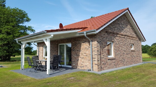 A small brick house with a red roof, a covered patio with a table and chairs, and a brick exterior.
