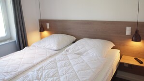 A double bed with a wooden headboard, two bedside lamps, a remote control on a nightstand, and a window with curtains.