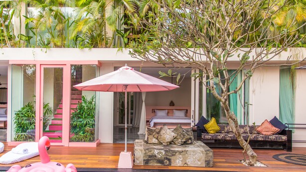 Ein Poolbereich mit einem rosa Flamingo-Schwimmpfad, eine Lounge mit einer Couch und Kissen sowie eine Treppe zu einem Balkon mit Pflanzen.