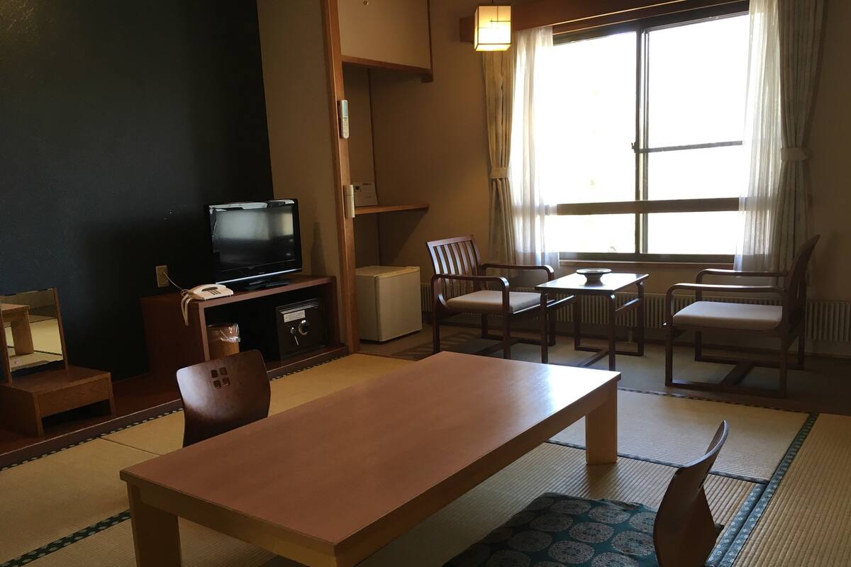 Une pièce de style japonais traditionnel, avec un sol recouvert de tatamis, une petite table basse, deux chaises, un téléviseur et un petit meuble.