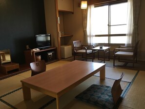 Una habitación tradicional japonesa con suelo de tatami, una mesa baja, dos sillas, un televisor y un armario pequeño.