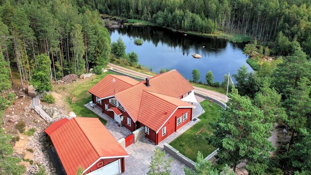 Ein Haus mit rotem Dach, einem gepflasterten Einfahrt, einer kleinen Rasenfläche und einem Teich, der von dichtem Wald umgeben ist.
