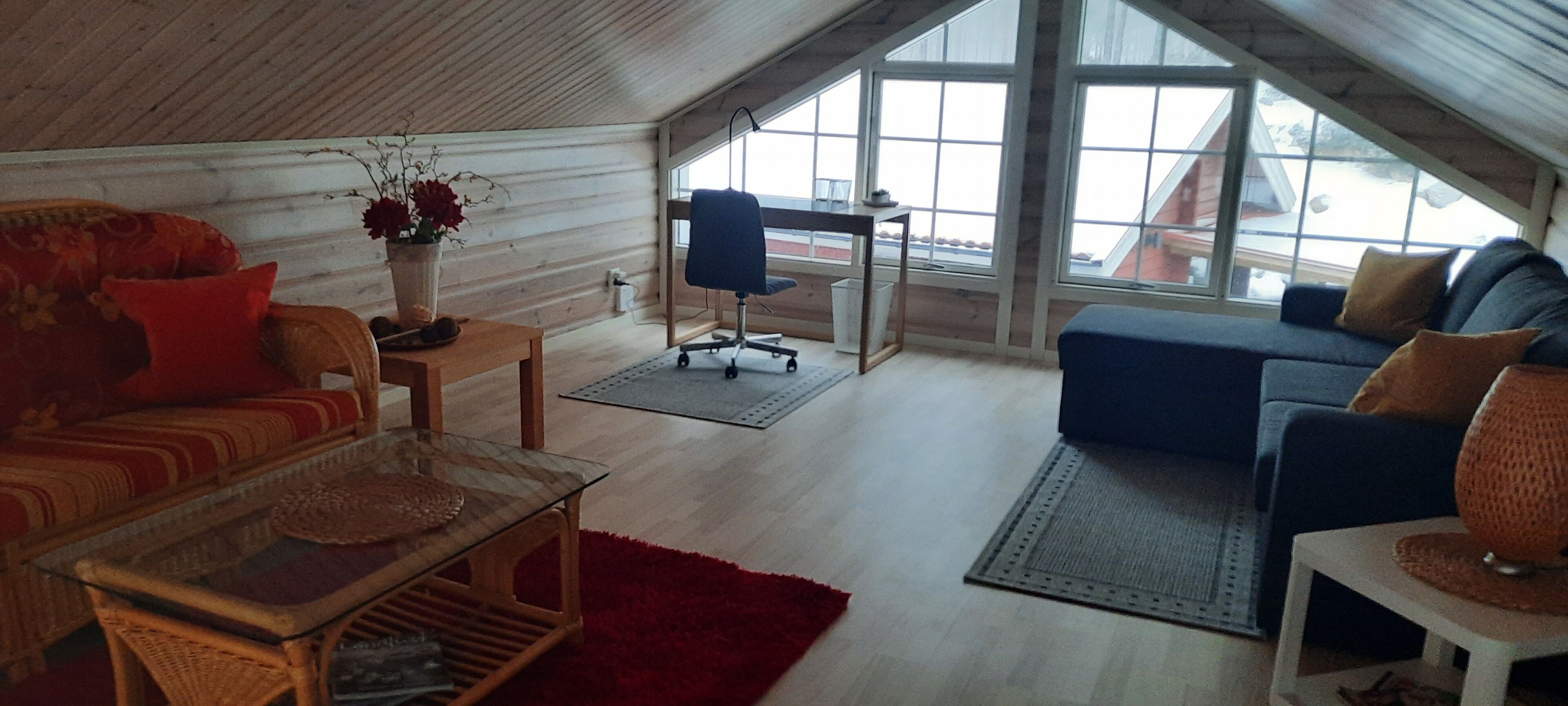 Um interior acolhedor com teto de madeira, um sofá, uma poltrona, uma mesa pequena e uma janela grande com vista para o exterior.