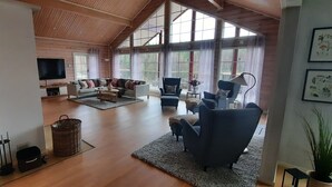 Ein geräumiges Wohnzimmer mit einer großen Eckcouch, einem Couchtisch und mehreren Sesseln. Ein Fernseher ist an der Wand befestigt, ein großes Fenster lässt Tageslicht herein und es gibt einen Kamin.