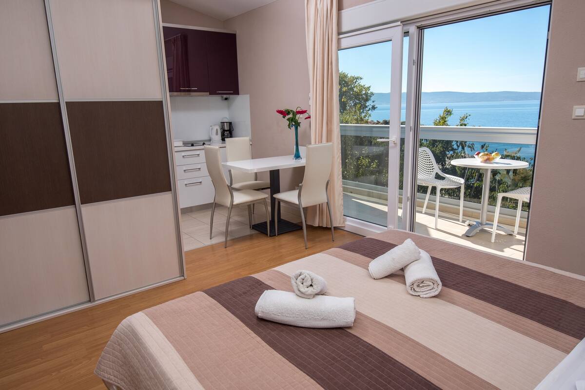 A hotel room with a bed, a desk, a chair, a small table with a vase of flowers, and a balcony with a view of the sea.