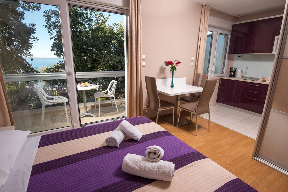 A hotel room with a bed, a desk, a chair, a small table, a vase with flowers, and a view of the sea.