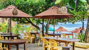 An outdoor seating area with wooden tables and yellow plastic chairs, surrounded by large umbrellas and lush greenery.