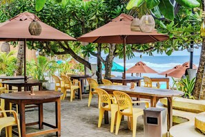 An outdoor seating area with wooden tables and yellow plastic chairs, surrounded by large umbrellas and lush greenery.