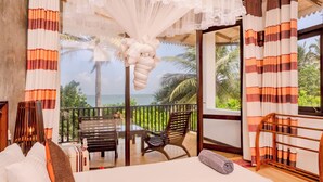 A bedroom with a bed, pillows, a wooden chair, a bench, and a view of the ocean.