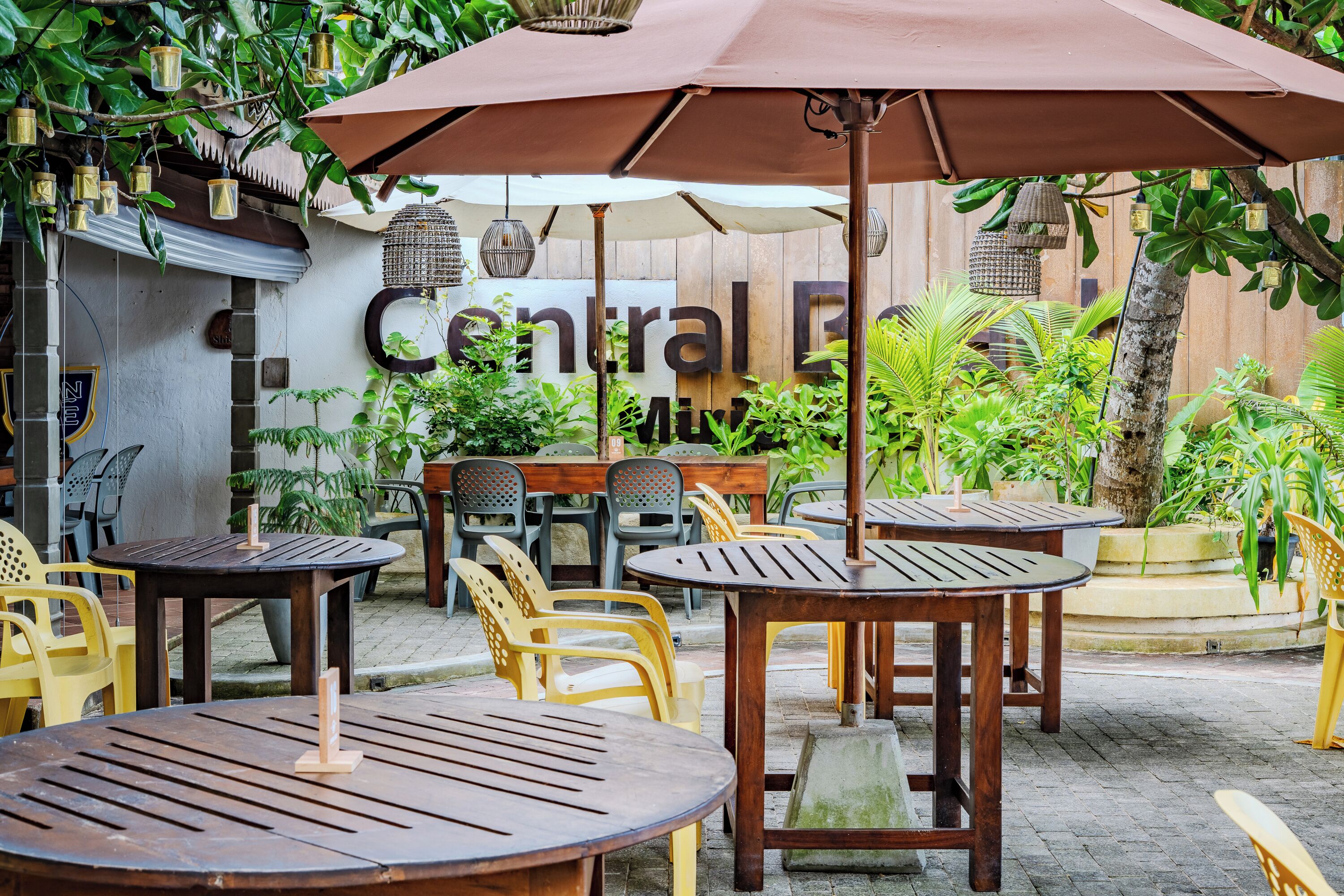 An outdoor seating area with wooden tables and chairs, surrounded by greenery and a wooden fence.