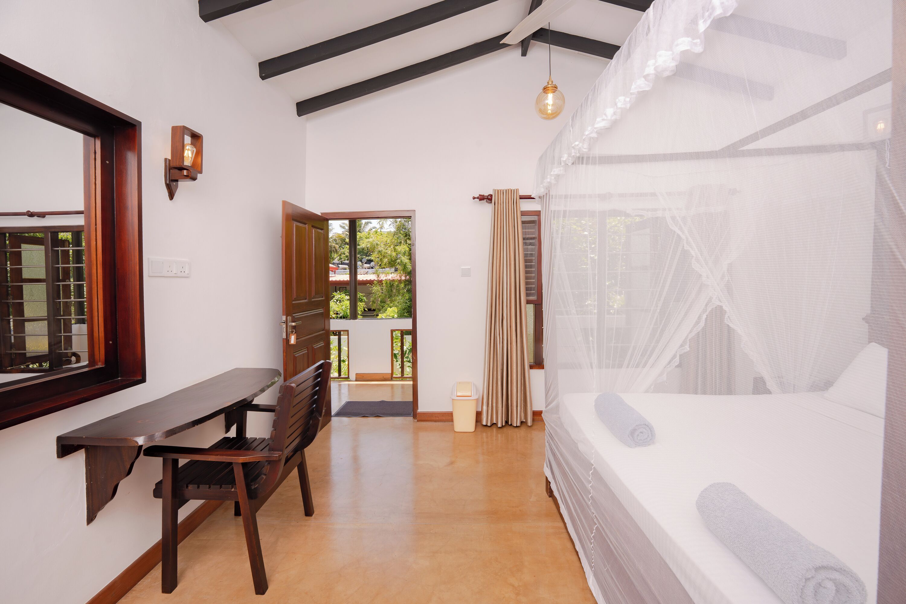 A bedroom with a bed, a desk, and a chair. There is a window with curtains and a view of greenery outside.